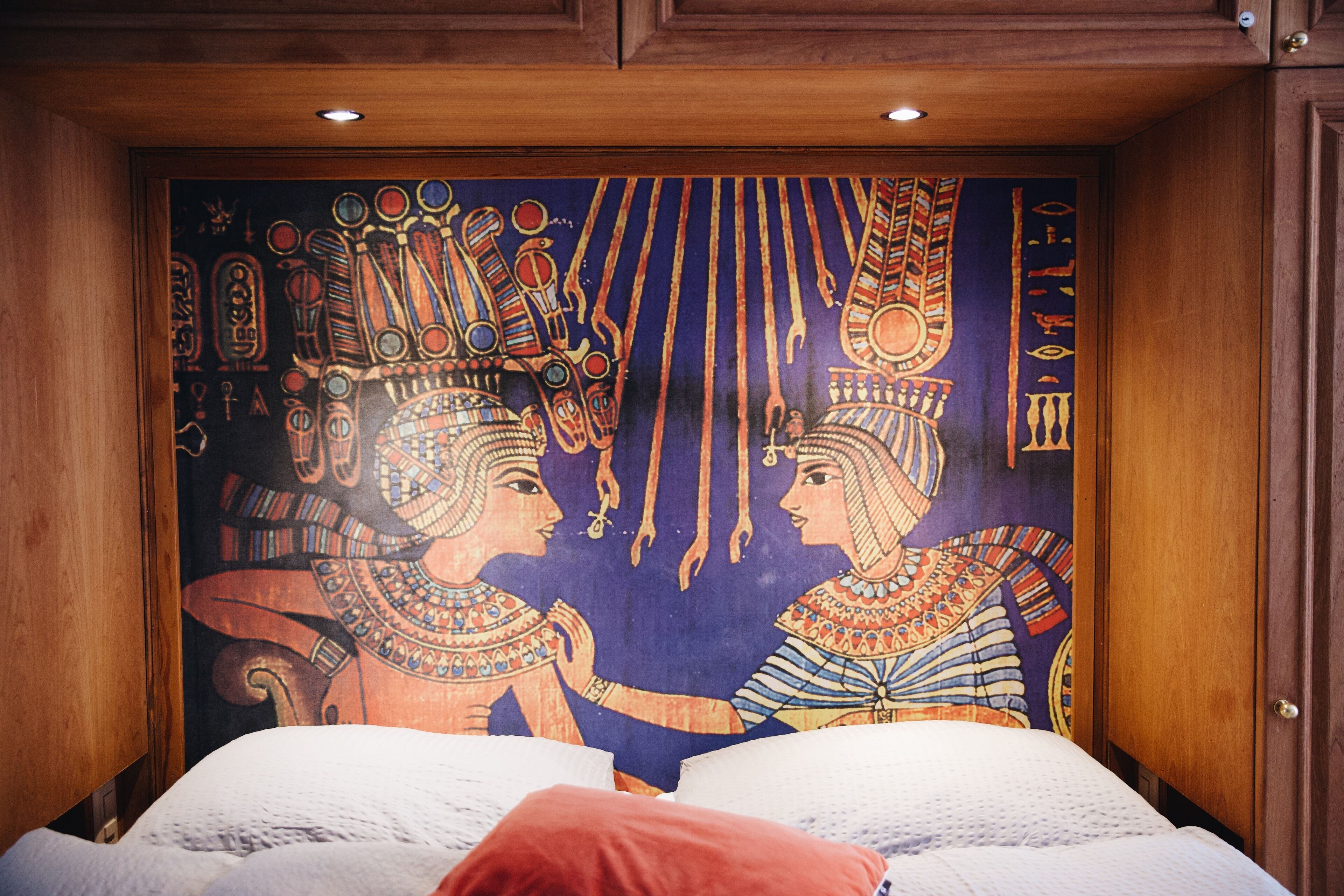 Ein Zimmer mit einer großen Wandmalerei von altägyptischen Figuren, einem hölzernen Kopfteil und einem roten Kissen.