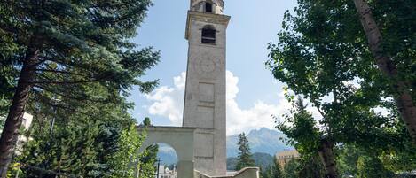 A tall clock tower surrounded by trees and greenery, with mountains in the background.