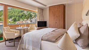 A modern hotel room with a large bed, a television, a dining table with chairs, and a view of a mountainous landscape. - Hotel Arte (St. Moritz)