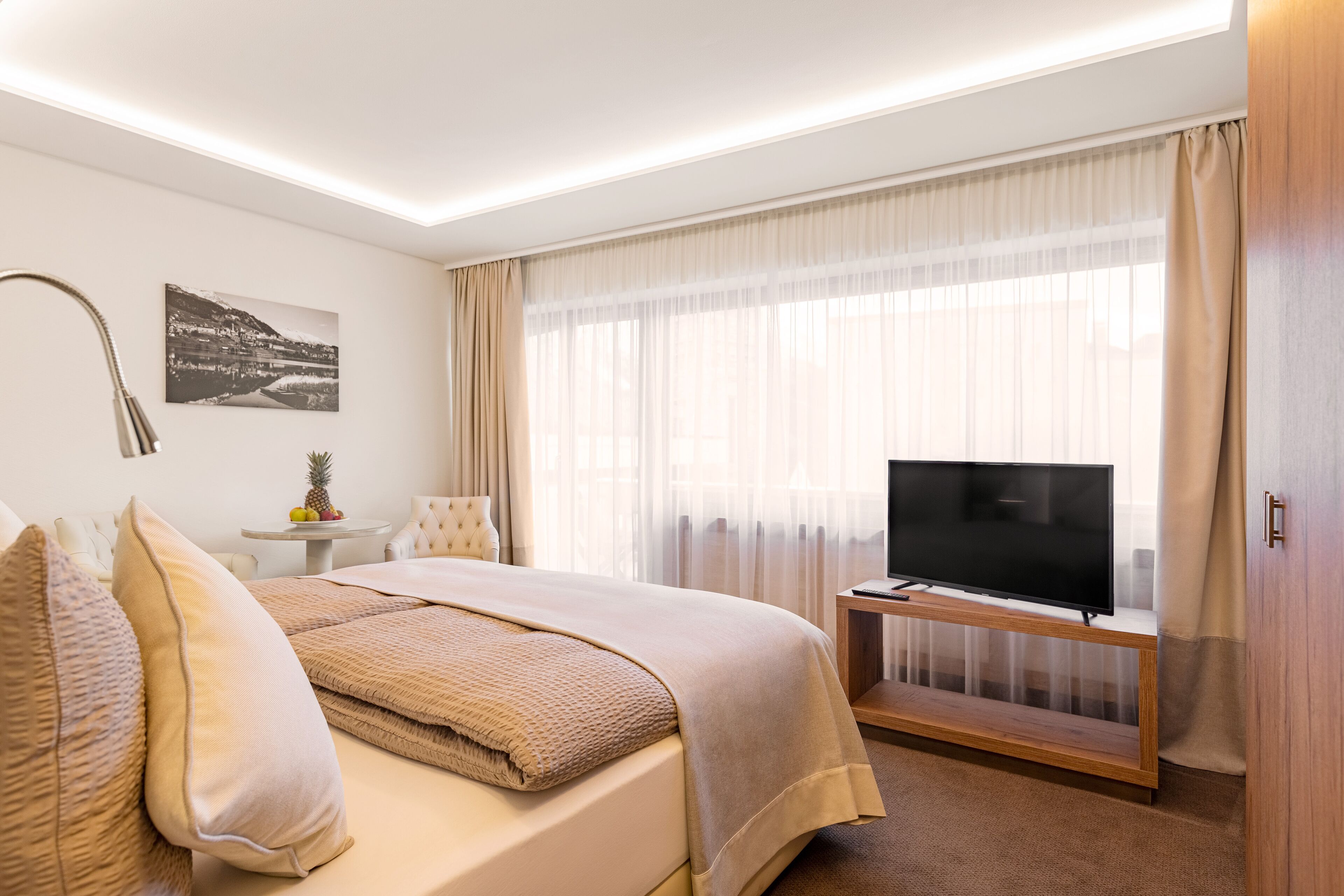 Quarto de hotel com uma cama, uma televisão sobre um suporte de madeira, uma mesinha com frutas e um quadro na parede.