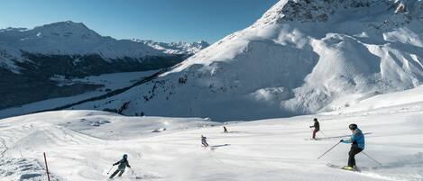 A snowy ski slope with skiers, a clear sky, and snow-covered mountains in the background.