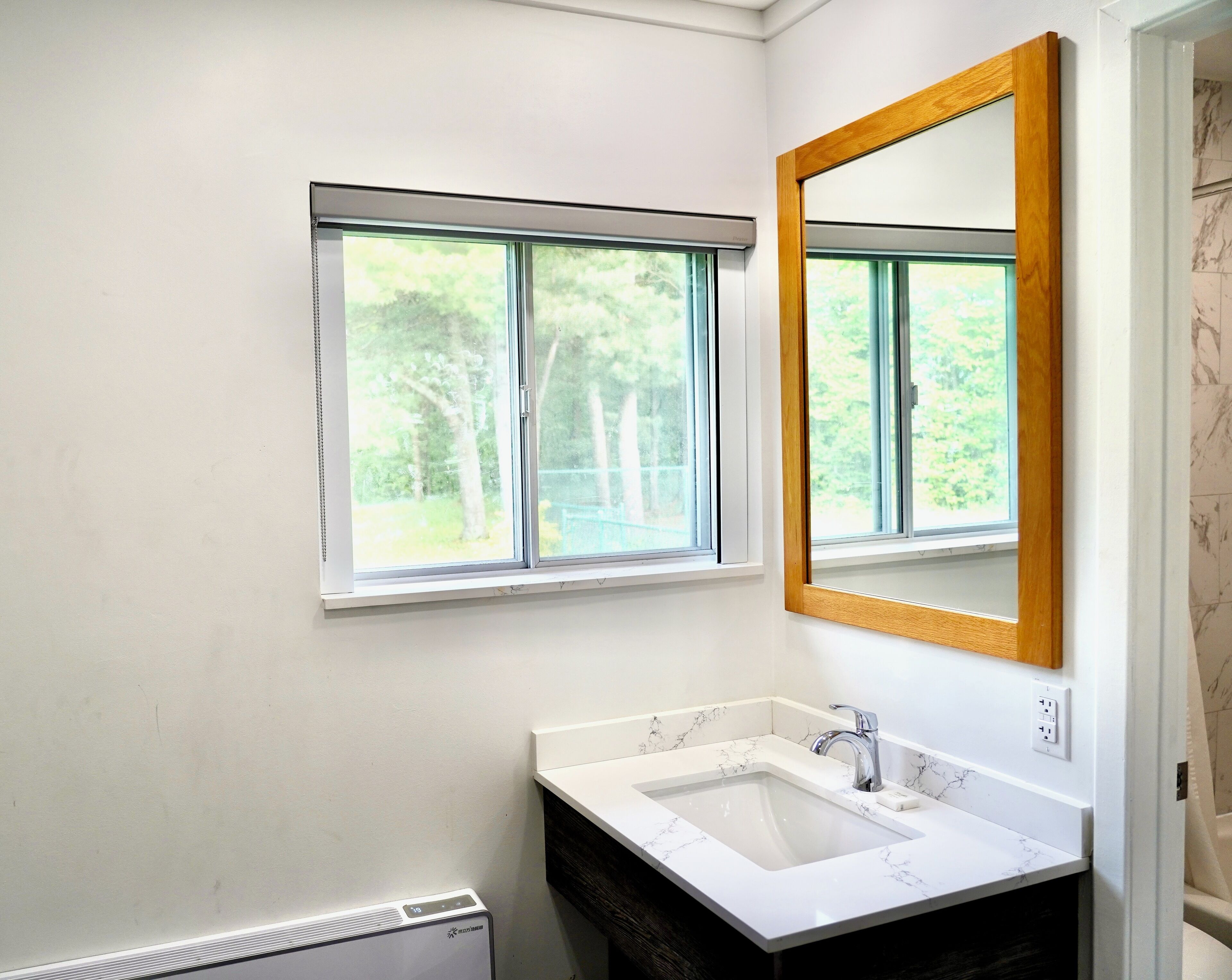A bathroom with a white sink, a wooden-framed mirror, and a window with a view of trees.