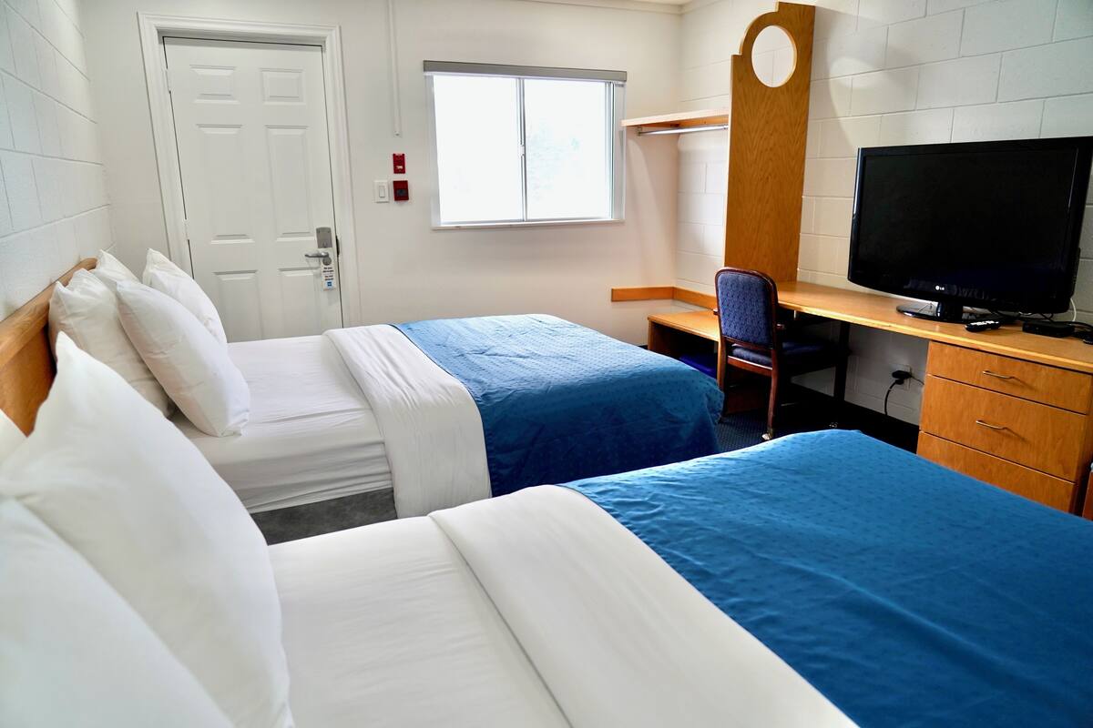 Quarto de hotel com duas camas, uma escrivaninha com televisão, uma cadeira e um guarda-roupa.