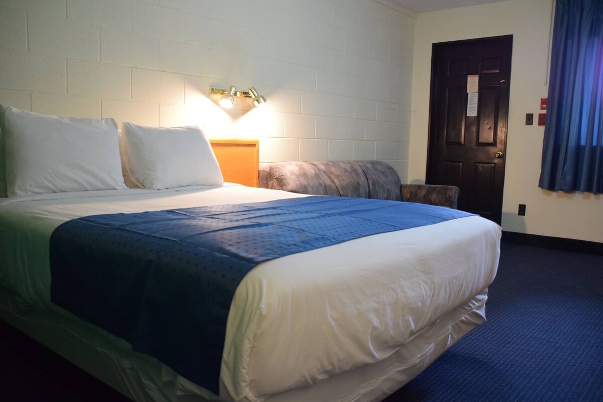 Quarto de hotel com uma cama grande, uma mesa de cabeceira, uma luminária fixada na parede e uma porta com cortina azul.