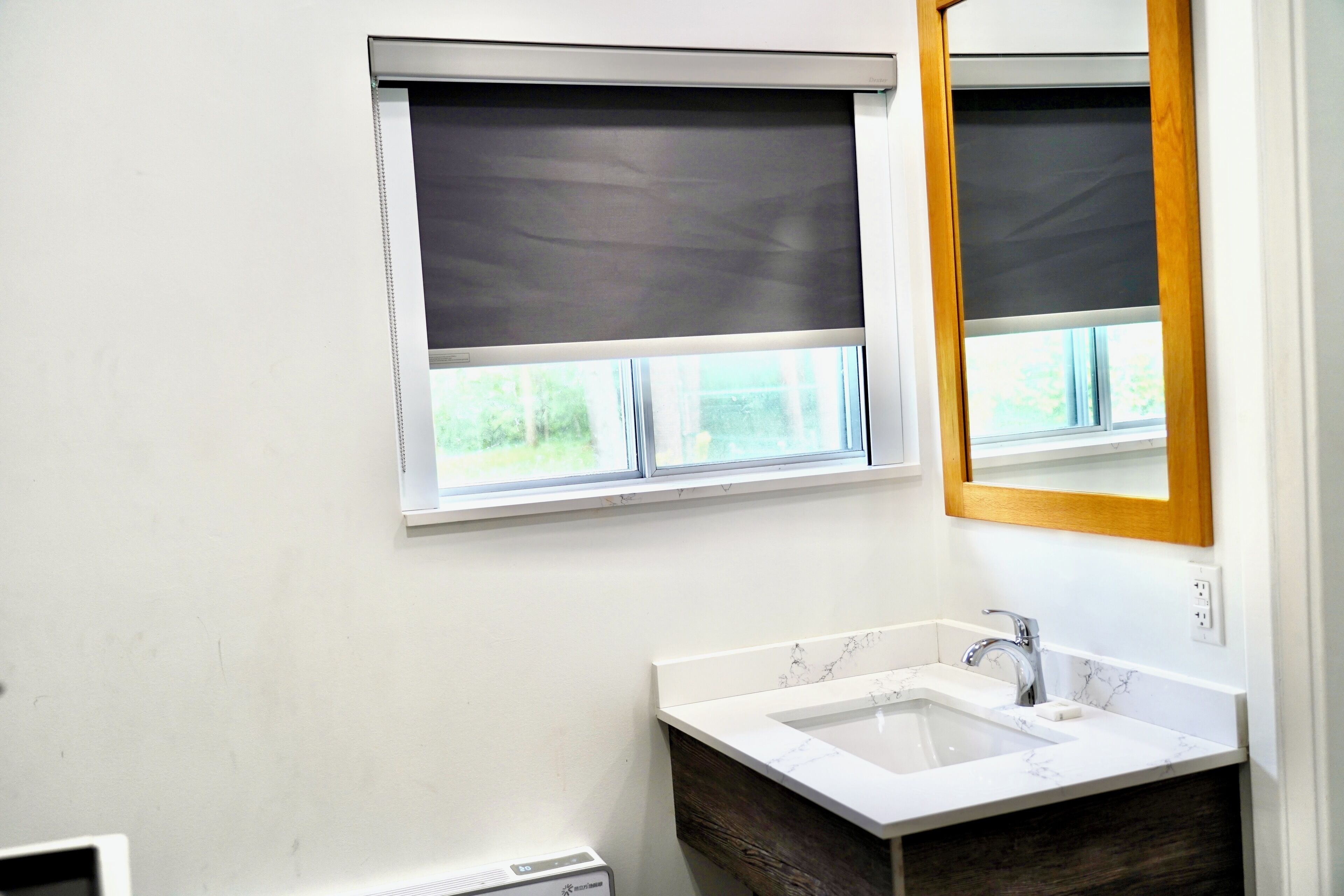 Une salle de bain avec un lavabo blanc, un meuble-lavabo en bois et deux fenêtres équipées de stores.