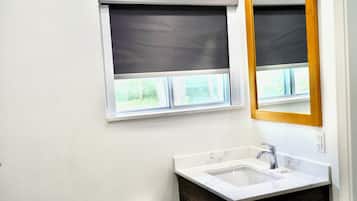 Une salle de bain avec un lavabo blanc, un meuble-lavabo en bois et deux fenêtres équipées de stores.