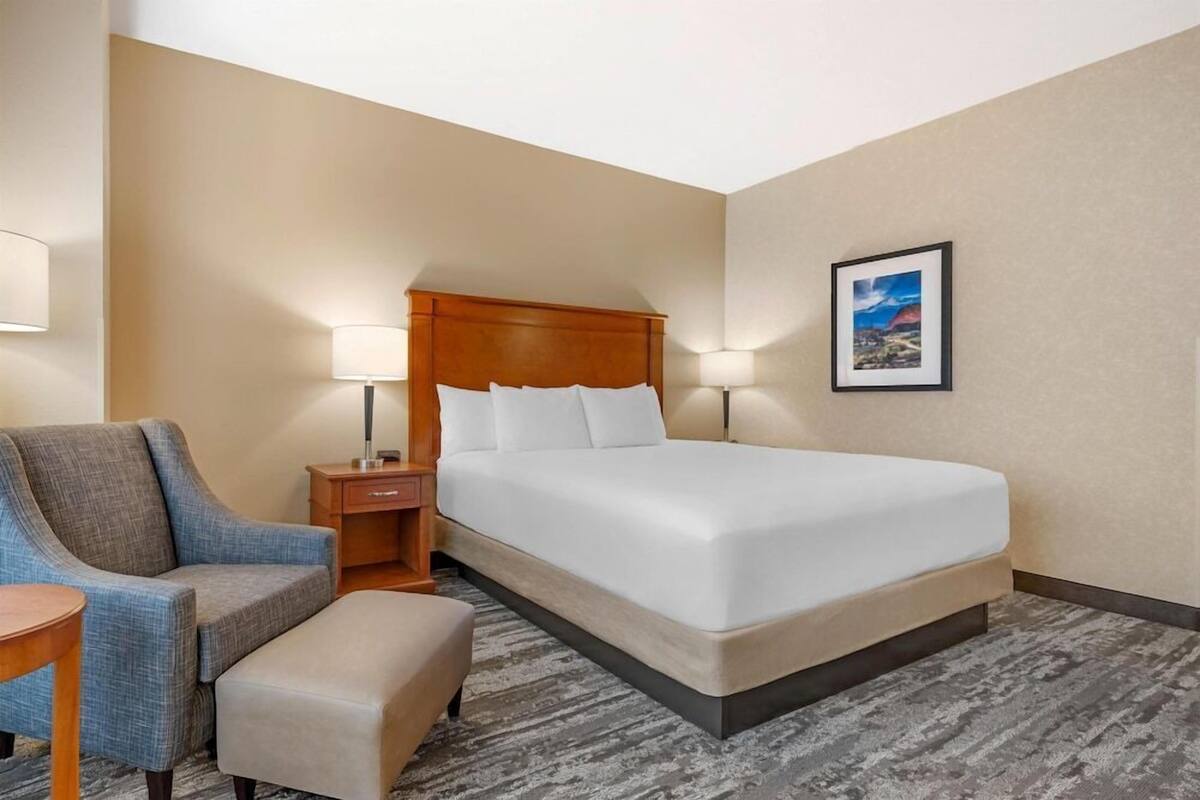 Une chambre d’hôtel avec un lit, une chaise, une table de chevet, une lampe et un tableau encadré au mur.