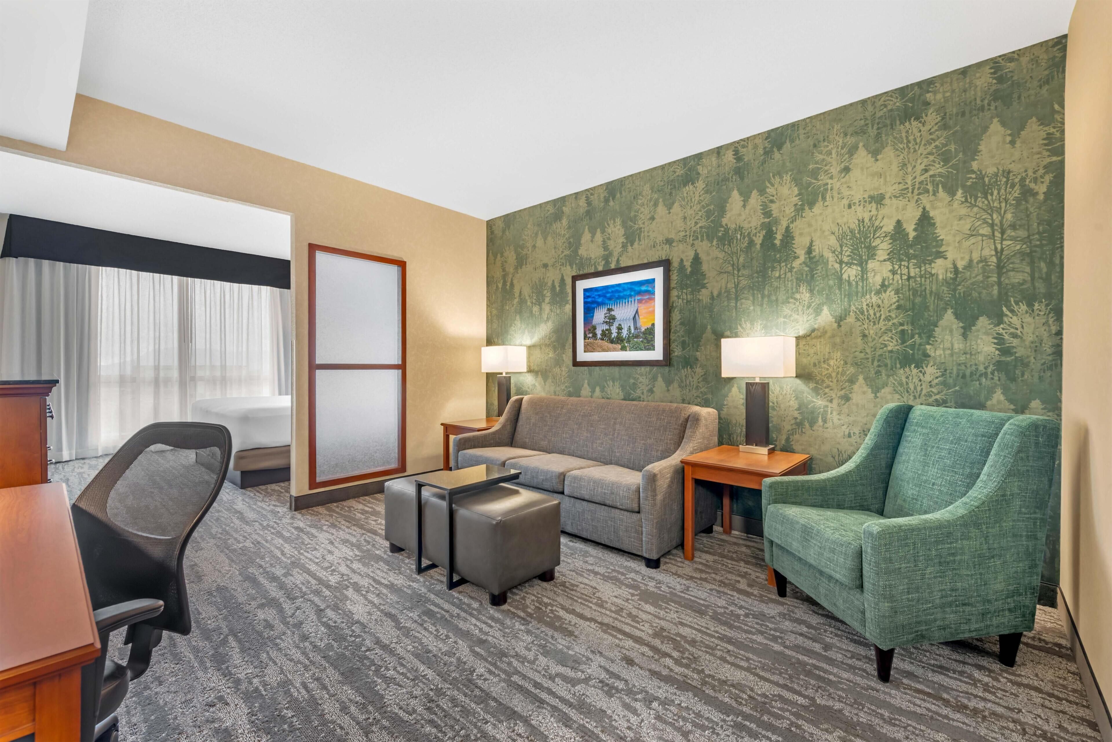 A hotel room with a gray carpet, a gray sofa, a green armchair, a wooden desk, a black office chair, and a framed picture on the wall.