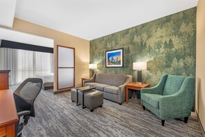 Une chambre d’hôtel avec un tapis gris, un canapé gris, un fauteuil vert, un bureau en bois, une chaise de bureau noire et un tableau encadré au mur.