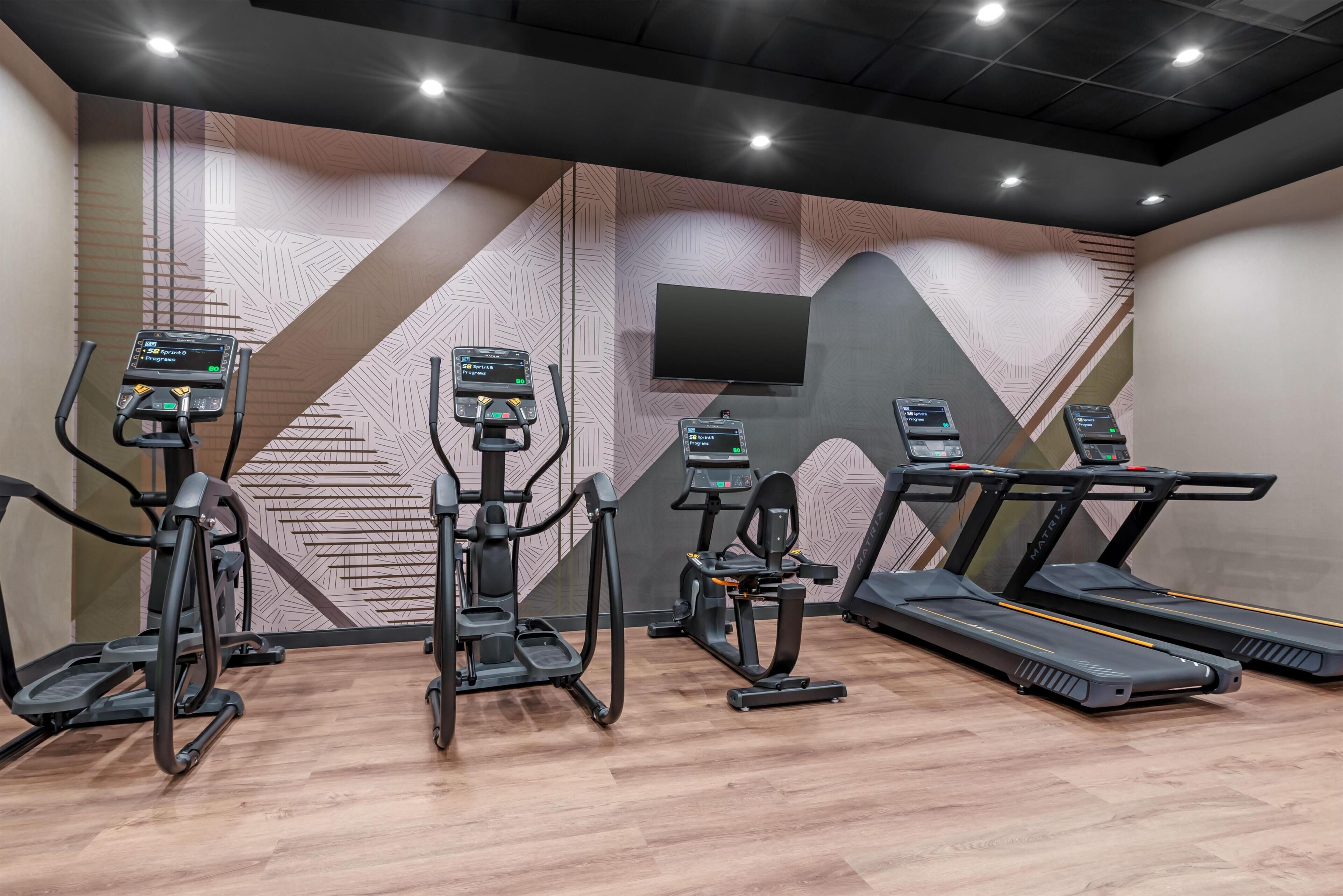 Un gimnasio con cintas de correr, elípticas y una bicicleta estática, con patrones geométricos en las paredes y un televisor montado.