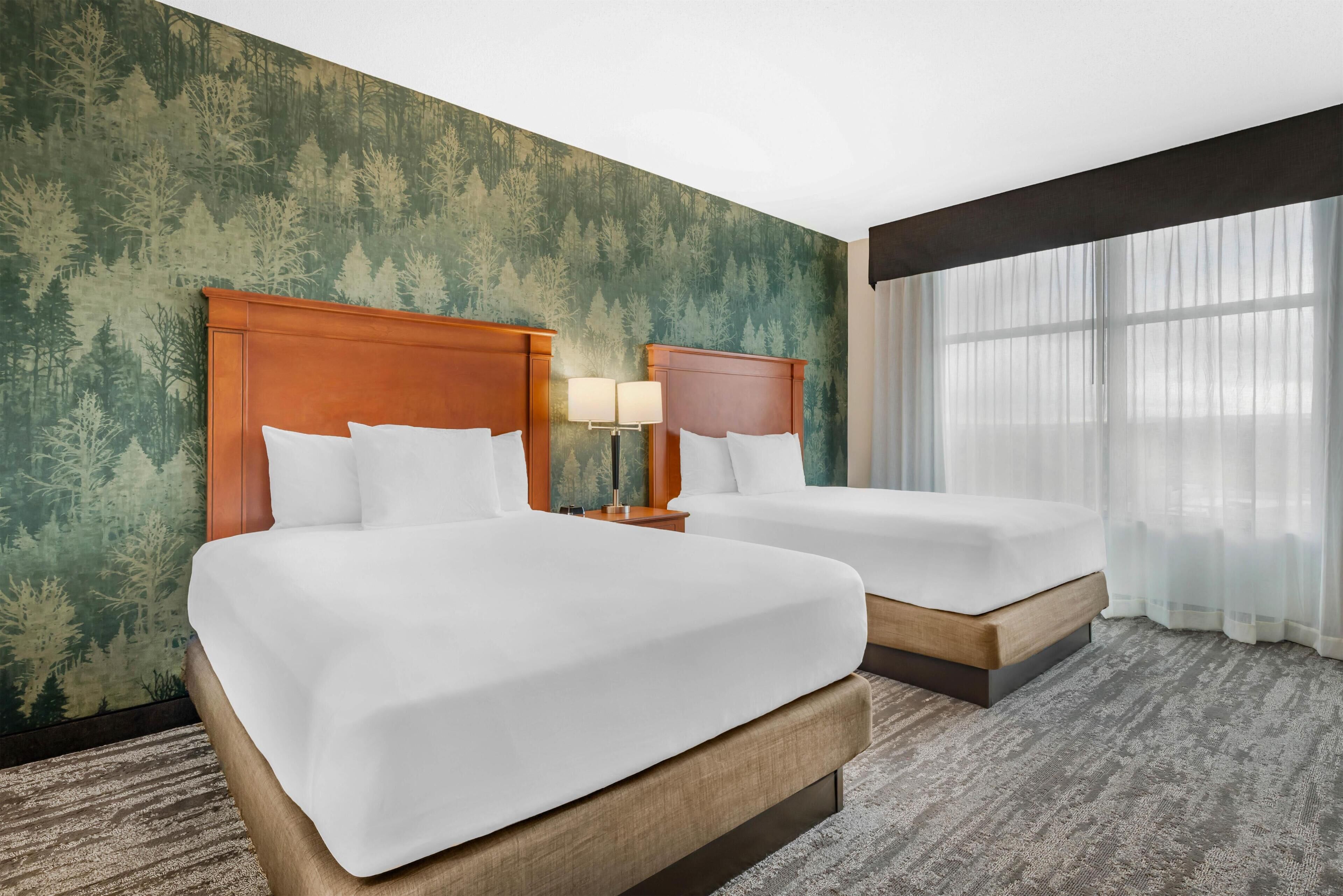 Two beds with wooden headboards, a nightstand with lamps, a large window with sheer curtains, and a forest-themed wall.