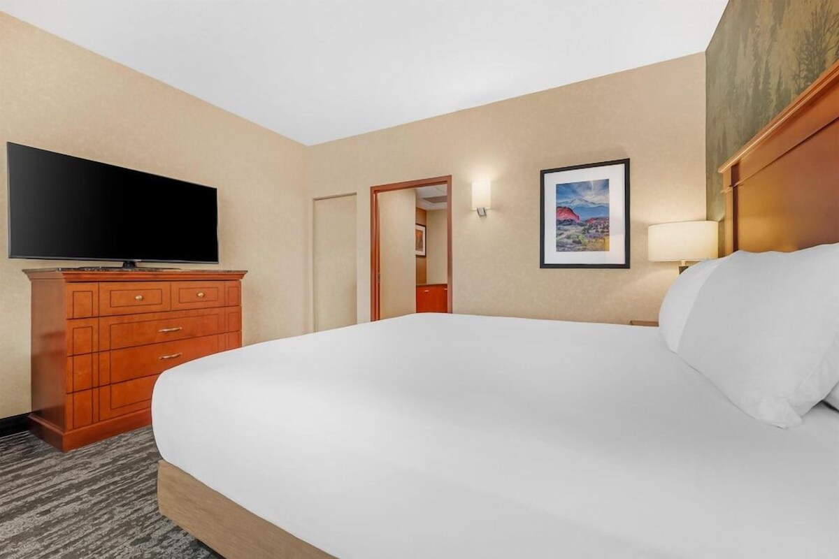 Une chambre d’hôtel avec un grand lit, une télévision, une commode en bois, un tableau encadré au mur et une porte donnant sur une autre pièce.