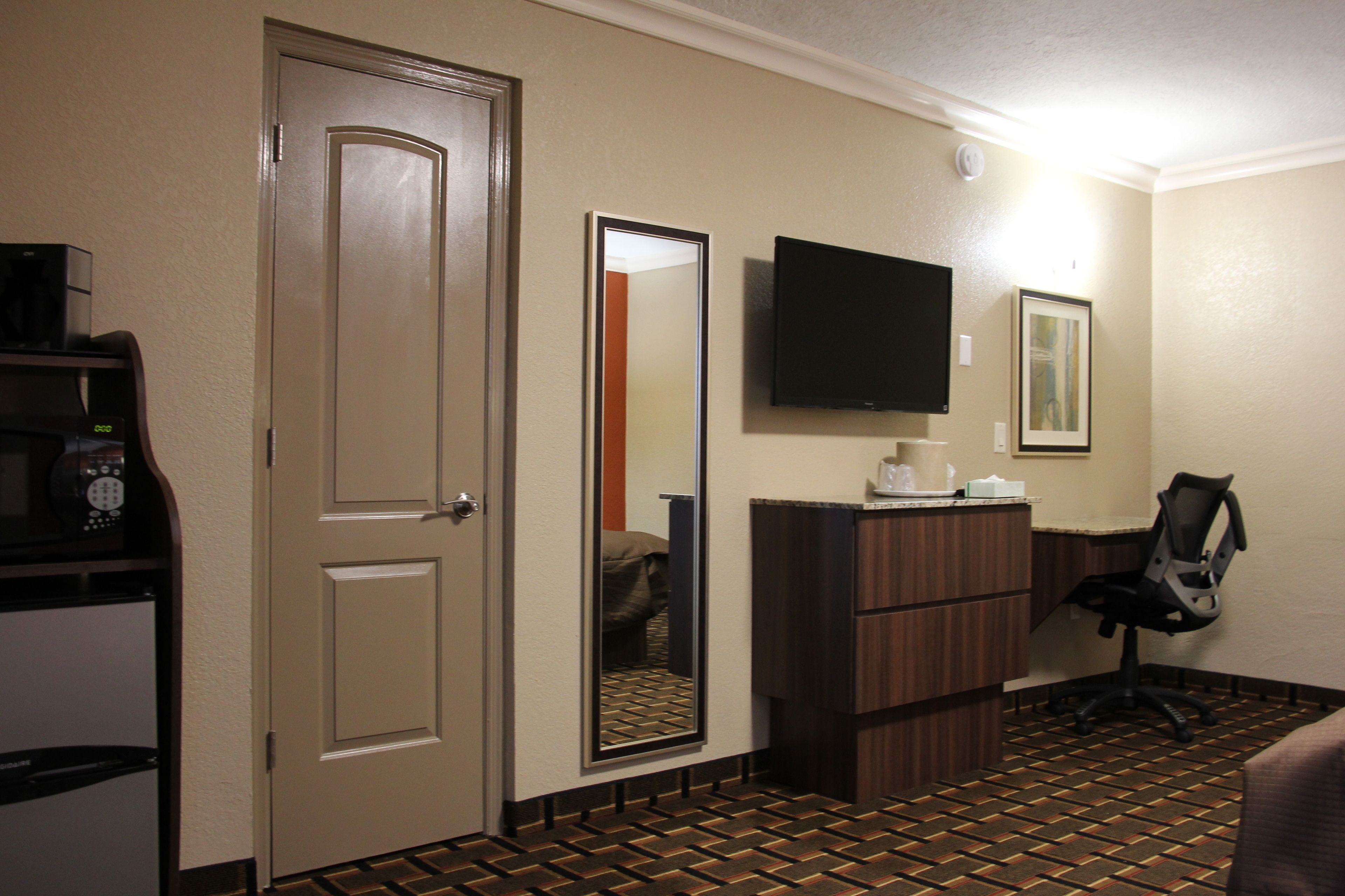 A hotel room with a door, a mirror, a flat-screen TV, a desk with a chair, and a patterned floor.