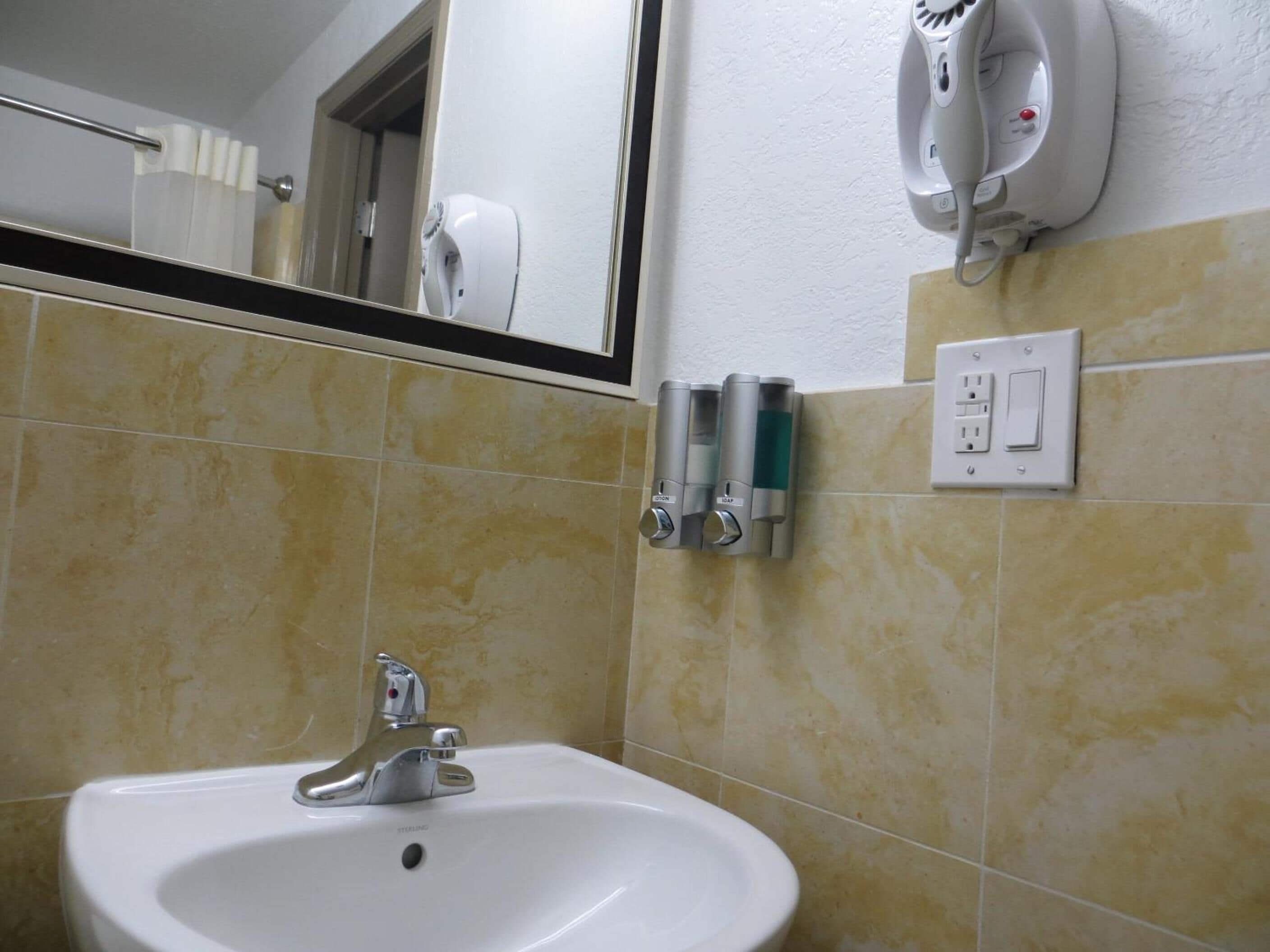 A bathroom with a sink, mirror, hand dryer, and soap dispensers.