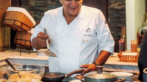 Een kok in een witte uniform met naamplaatje, kookt aan een keukenwerkblad met diverse keukengerei en ingrediënten.