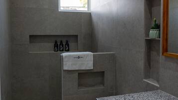 Un baño moderno con inodoro, lavamanos y zona de ducha. Hay artículos de aseo en un estante y una toalla colgada en la pared.
