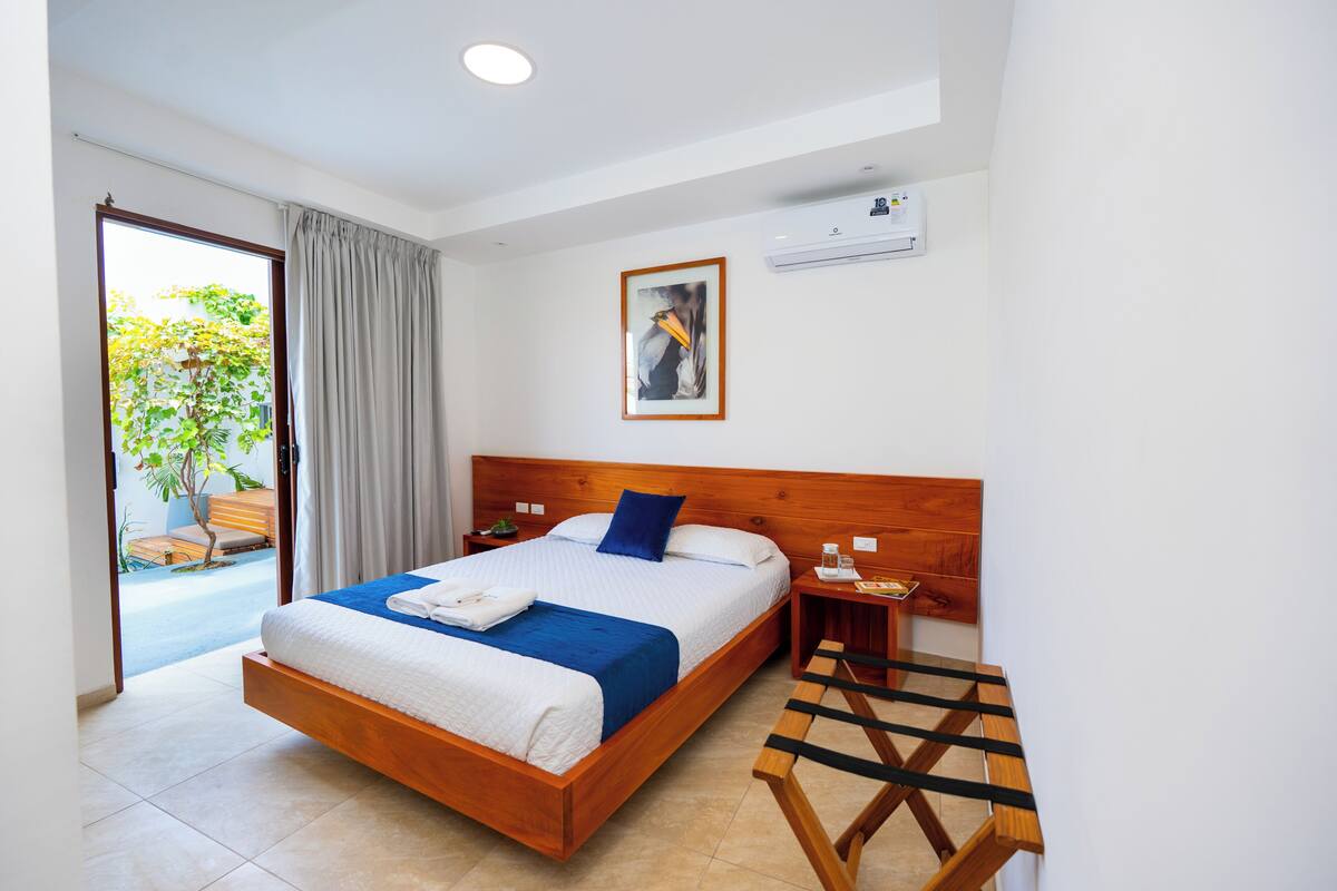 Une chambre d’hôtel avec un lit en bois, une table de chevet, une petite table, une fenêtre avec des rideaux et un appareil de climatisation.