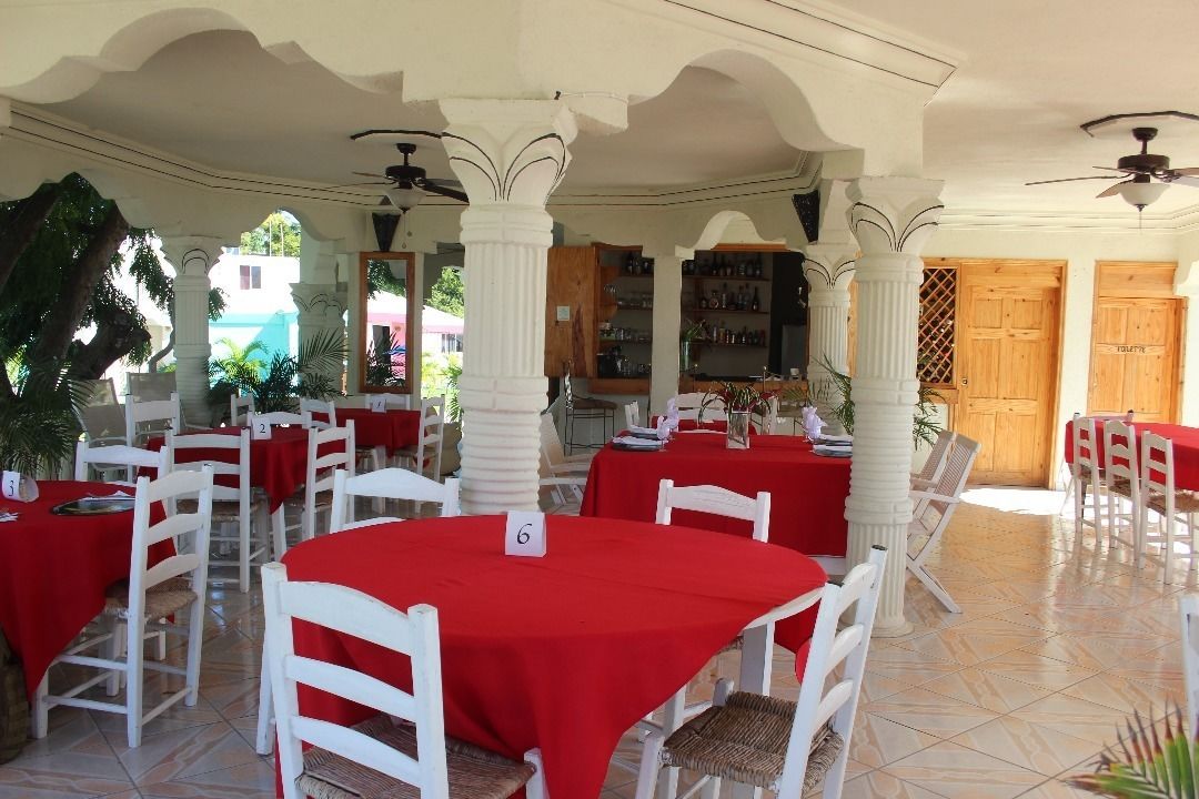 Een restaurant met witte stoelen en rode tafelkleden, met een betegelde vloer en zuilen.