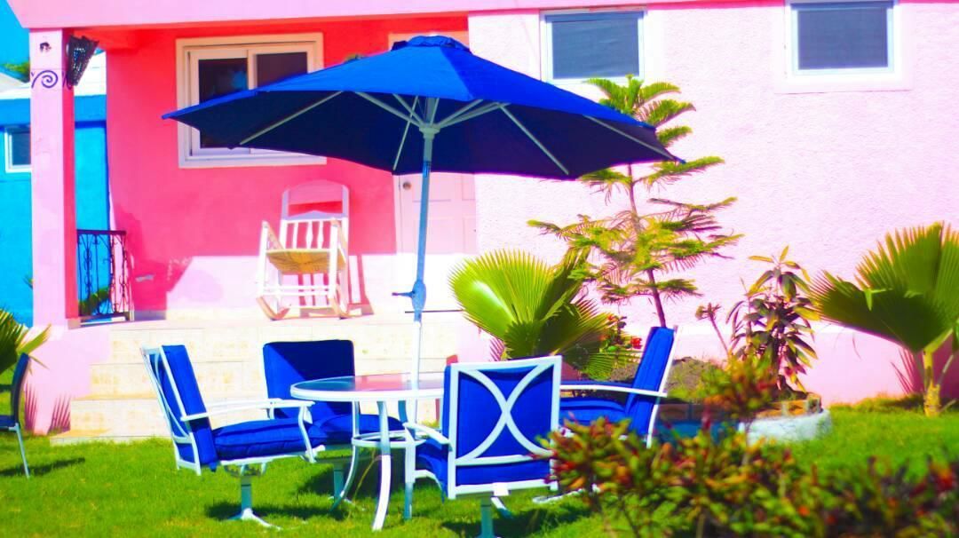 Un patio con sillas azules y una mesa, una sombrilla, y un edificio rosa con una puerta blanca.