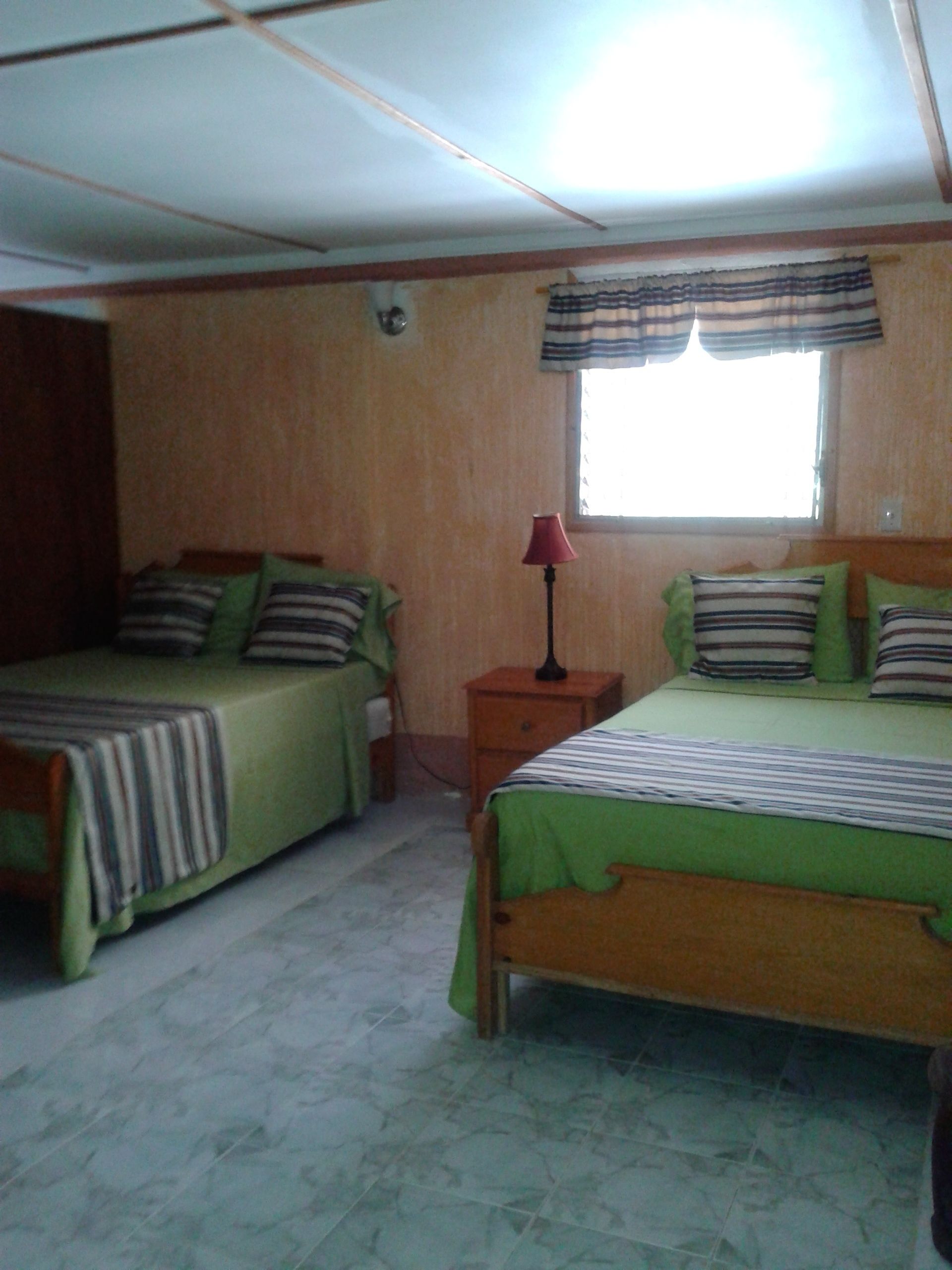 Una habitación con dos camas, cada una con ropa de cama y almohadas a rayas, una mesita de noche con una lámpara, y una ventana con cortina a rayas.