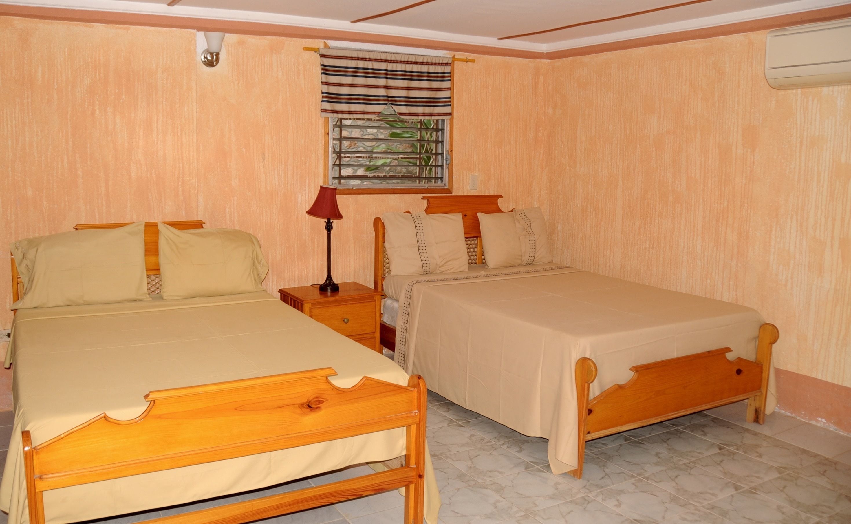 Un dormitorio con dos camas de madera, una mesita de noche con lámpara, una ventana con cortina a rayas y un aire acondicionado instalado en la pared.