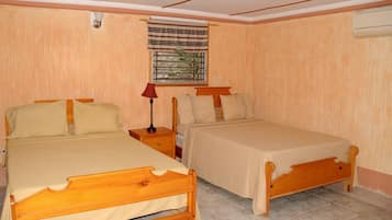Un dormitorio con dos camas de madera, una mesita de noche con lámpara, una ventana con cortina a rayas y un aire acondicionado instalado en la pared.