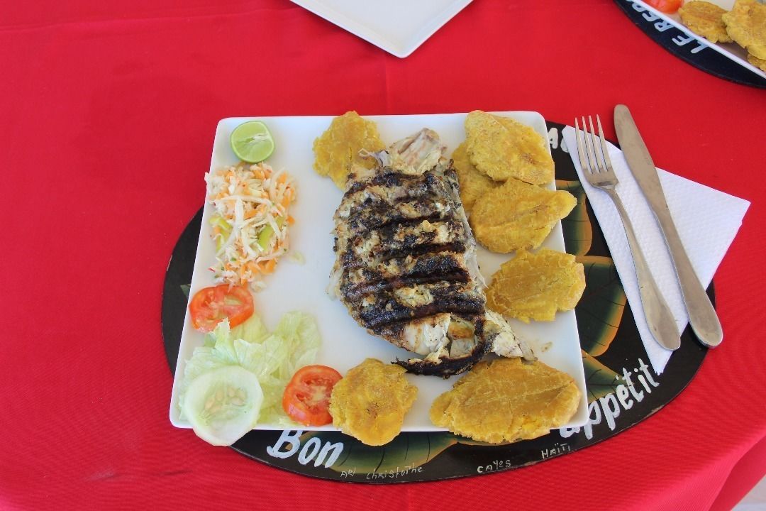 Um prato de peixe grelhado, banana frita e uma salada com um gajo de limão sobre uma mesa com toalha vermelha.