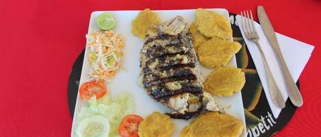 Um prato de peixe grelhado, banana frita e uma salada com um gajo de limão sobre uma mesa com toalha vermelha.