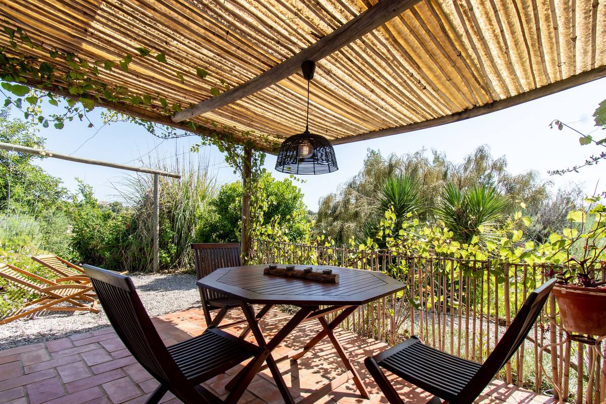 Une terrasse couverte avec une table ronde et des chaises, entourée de verdure et d’une clôture en bambou.