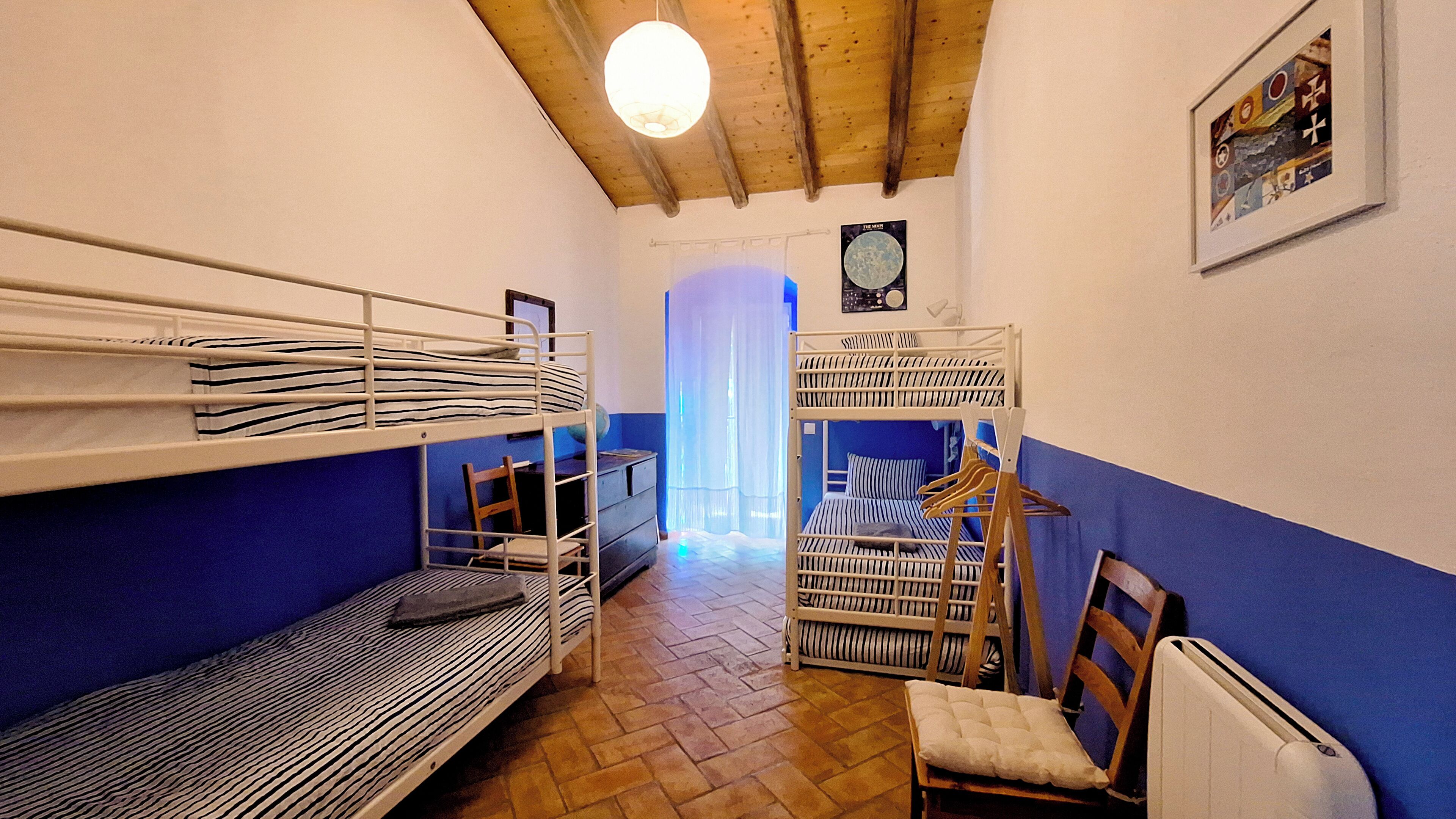 A dormitory room with bunk beds, a chair, a radiator, and a door leading to another room.