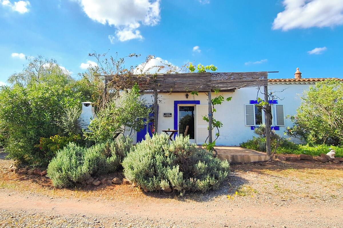 Un petit bâtiment avec une porte bleue et des volets aux fenêtres, entouré de verdure et bordé d’un chemin de gravier.