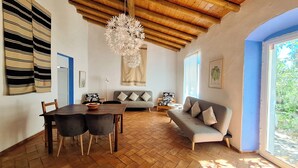 A dining area with a wooden table and chairs, a sofa with cushions, a hanging chandelier, and a view through a window. - Quinta da Fornalha (Castro Marim)