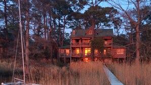 A wooden house with multiple balconies, surrounded by tall trees and a dock leading to a small boat. - Cypress Moon Inn (Kitty Hawk)