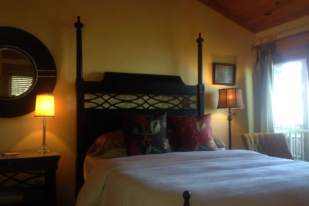 A four-poster bed with a wooden headboard, a bedside table with a lamp, a mirror on the wall, and a window with curtains.