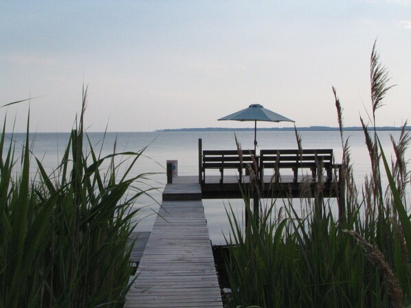 Une jetée en bois menant à un banc équipé d’un parasol, le tout entouré d’herbes hautes.