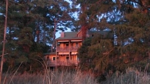 A two-story wooden cabin surrounded by tall grass and trees, with a visible pathway leading to the entrance. - Cypress Moon Inn (Kitty Hawk)