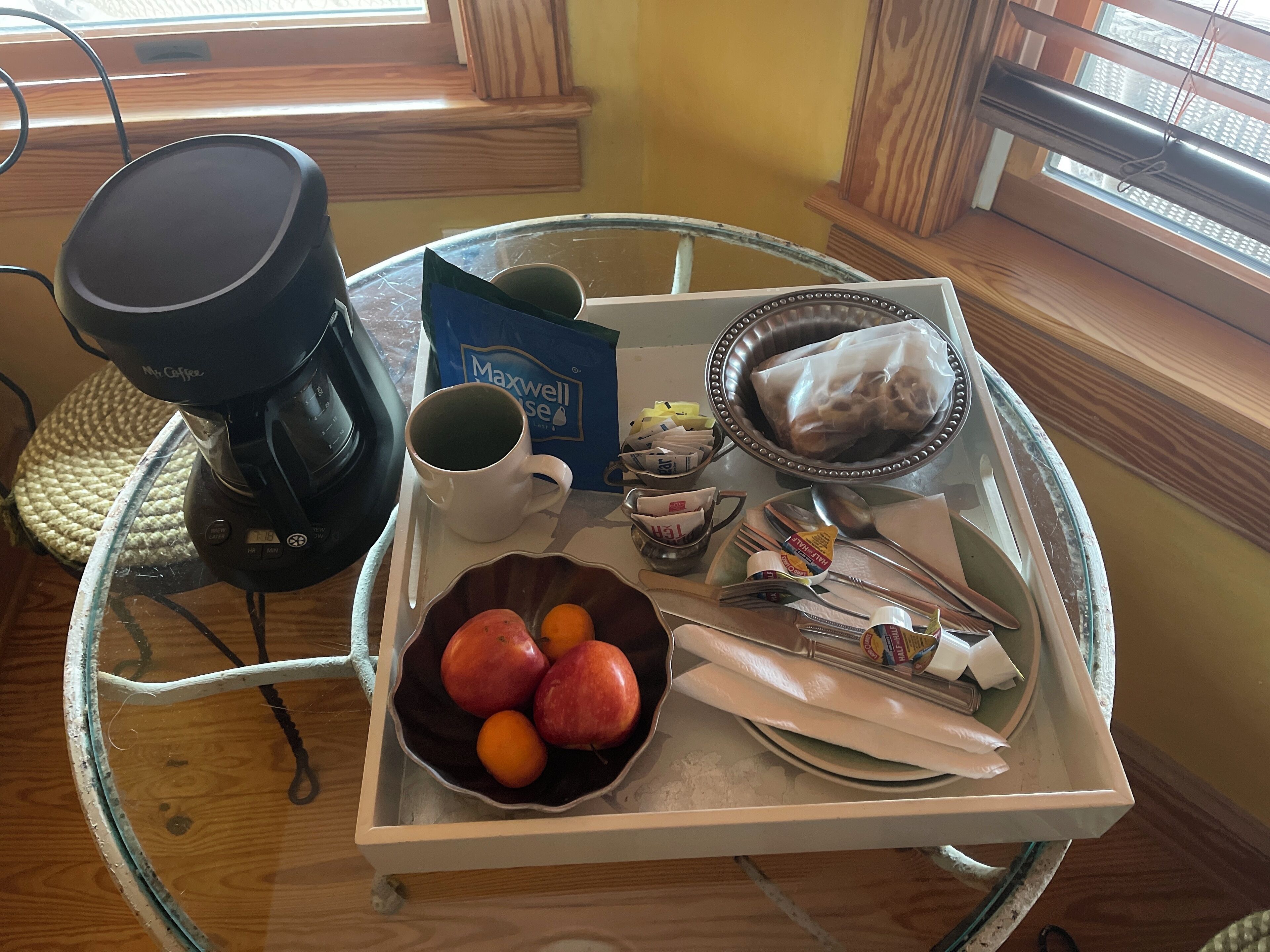 A breakfast tray with a coffee maker, a mug, a bowl of fruit, a Maxwell House coffee bag, and various breakfast items on a glass table.