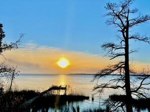 A sunset over a calm body of water with a pier and a leafless tree in the foreground.