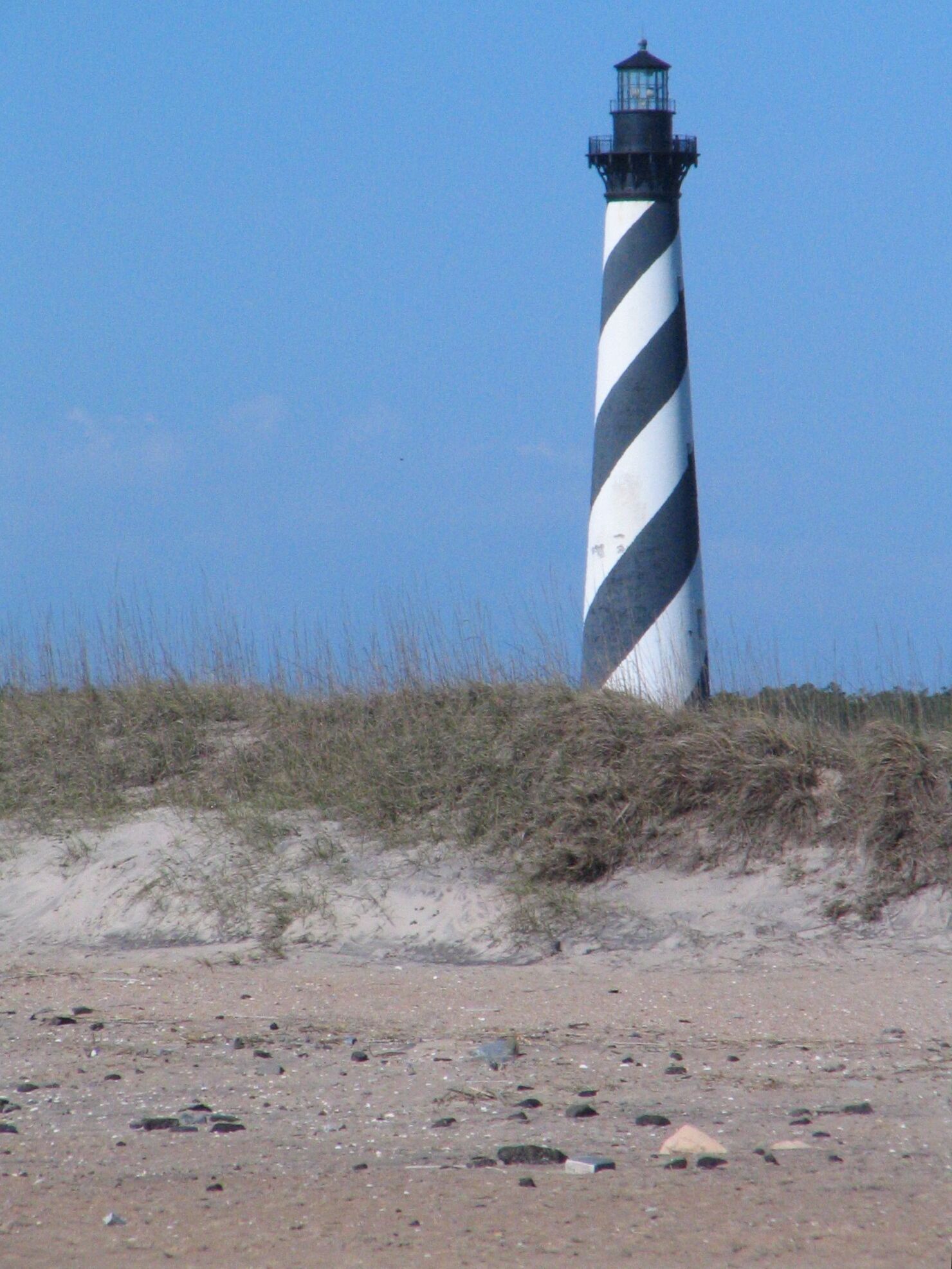 A lighthouse with a black and white striped pattern, situated on a sandy beach with sparse vegetation.