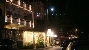 A building with a modern architectural design, featuring a balcony with lighting. There are cars parked in front, and a streetlight is visible. - Cyclades Hotel Serifos (Serifos)