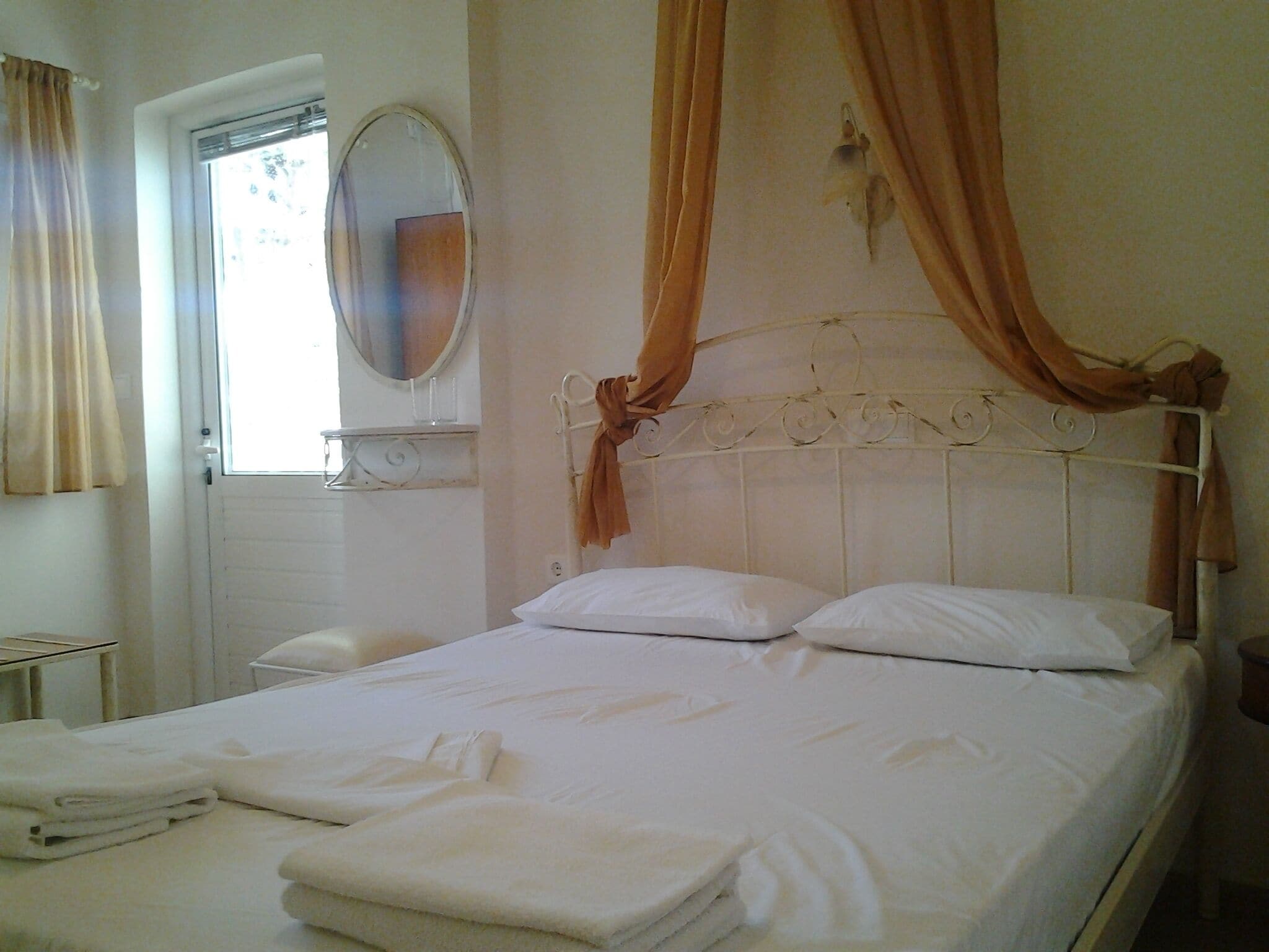 A neatly made bed with white linens and pillows, a bedside table, a mirror, and a window with curtains.