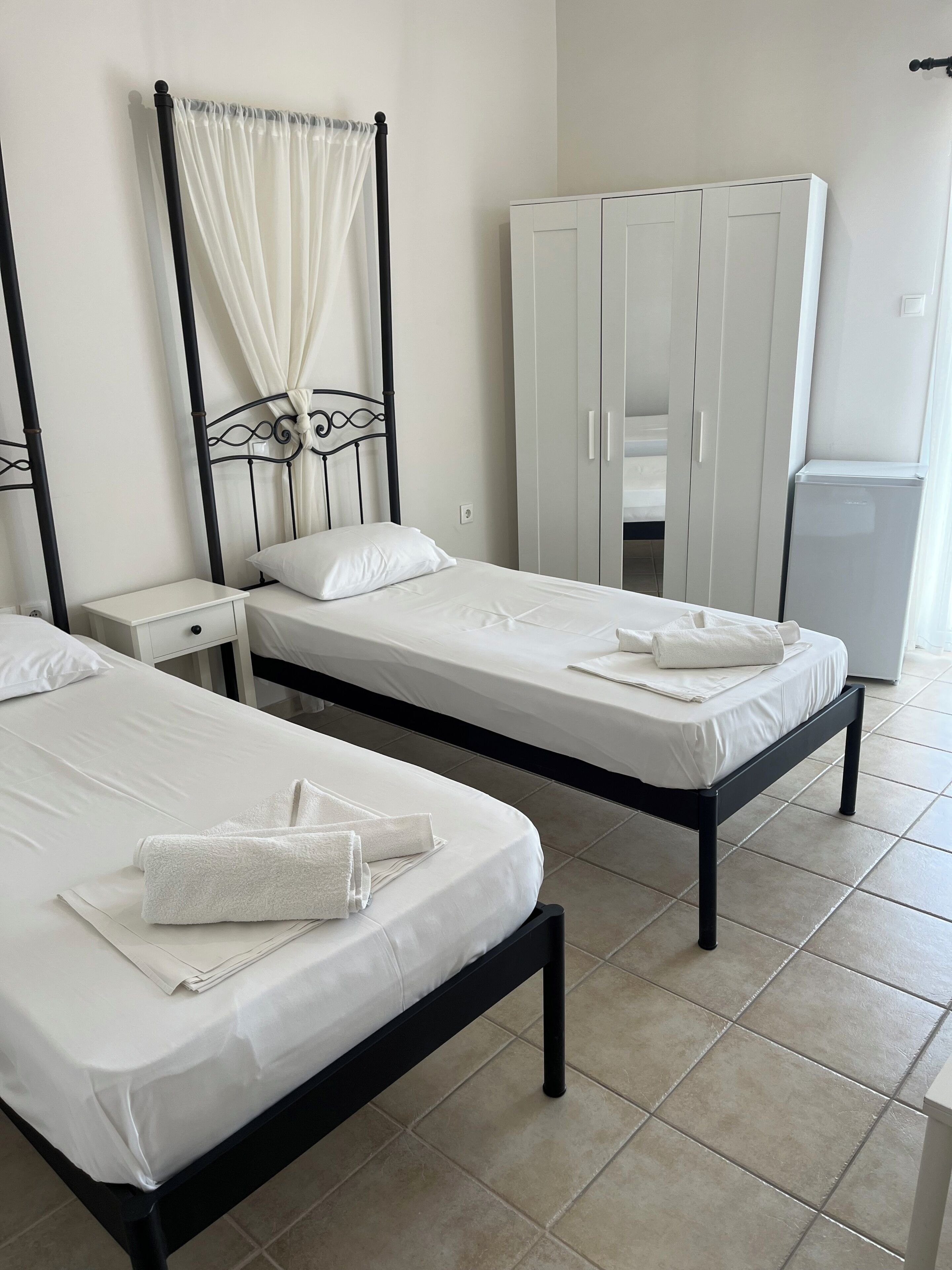 Two single beds with white linens, a wardrobe, and a small refrigerator in a room with tiled flooring.