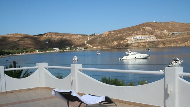 A balcony with a chair and a table, overlooking a calm body of water with boats and a hilly landscape in the background. - Cyclades Hotel Serifos (Serifos)