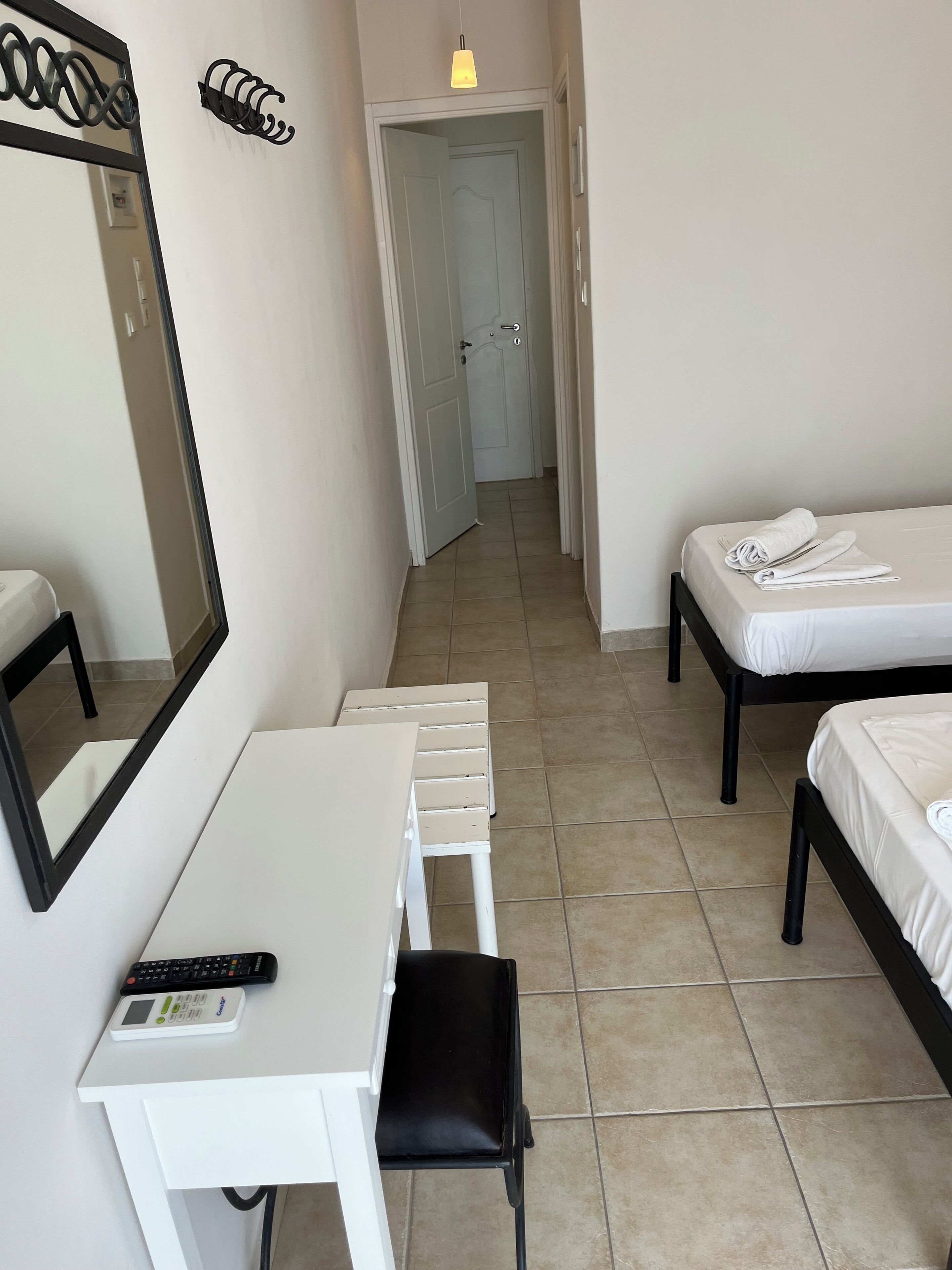 Quarto de hotel com uma cama, uma escrivaninha, uma cadeira, um espelho e uma porta que dá para outro espaço.