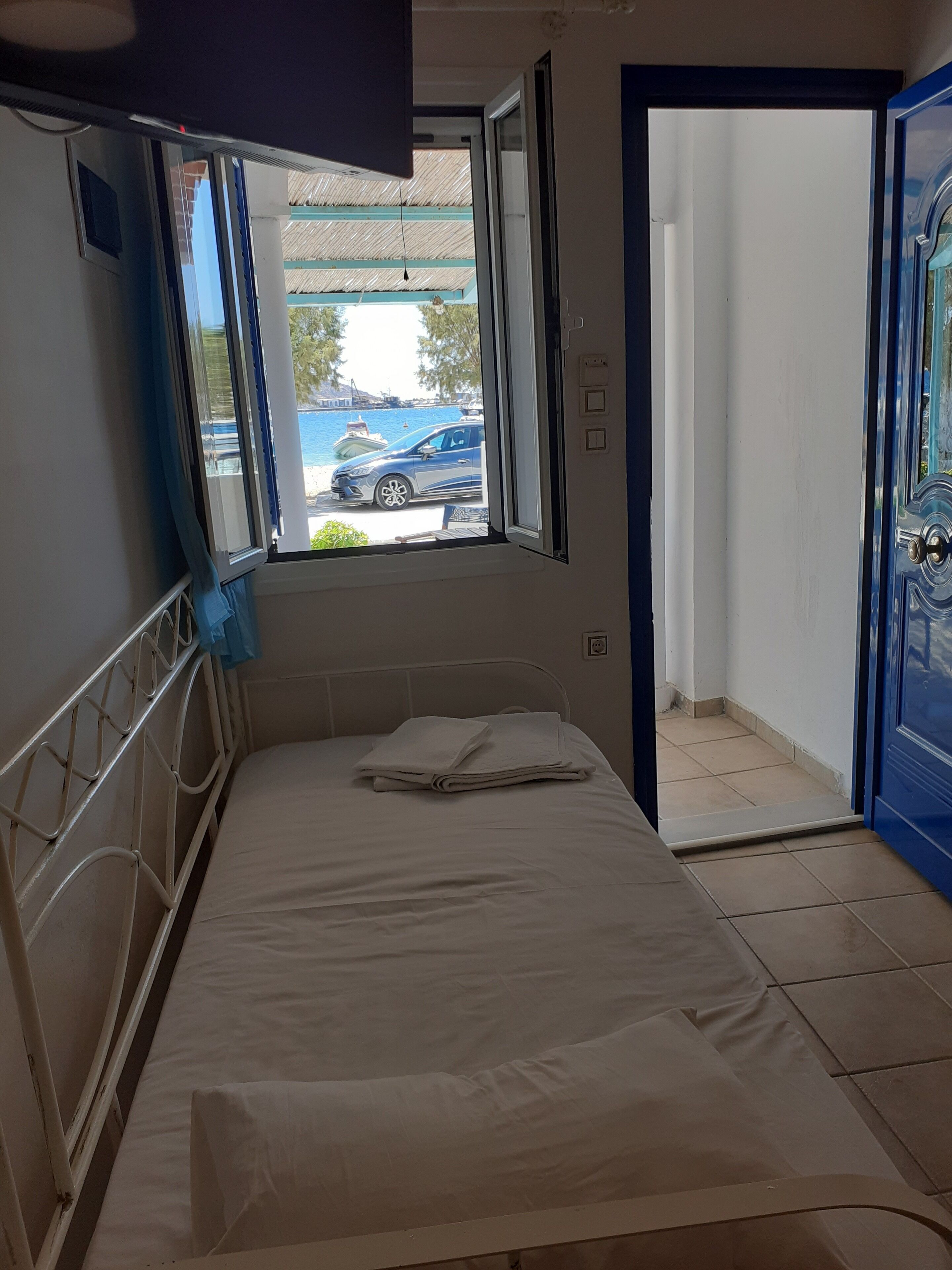 A single bed with a metal headboard in a room with a window, a door, and a view of a body of water and a car outside.