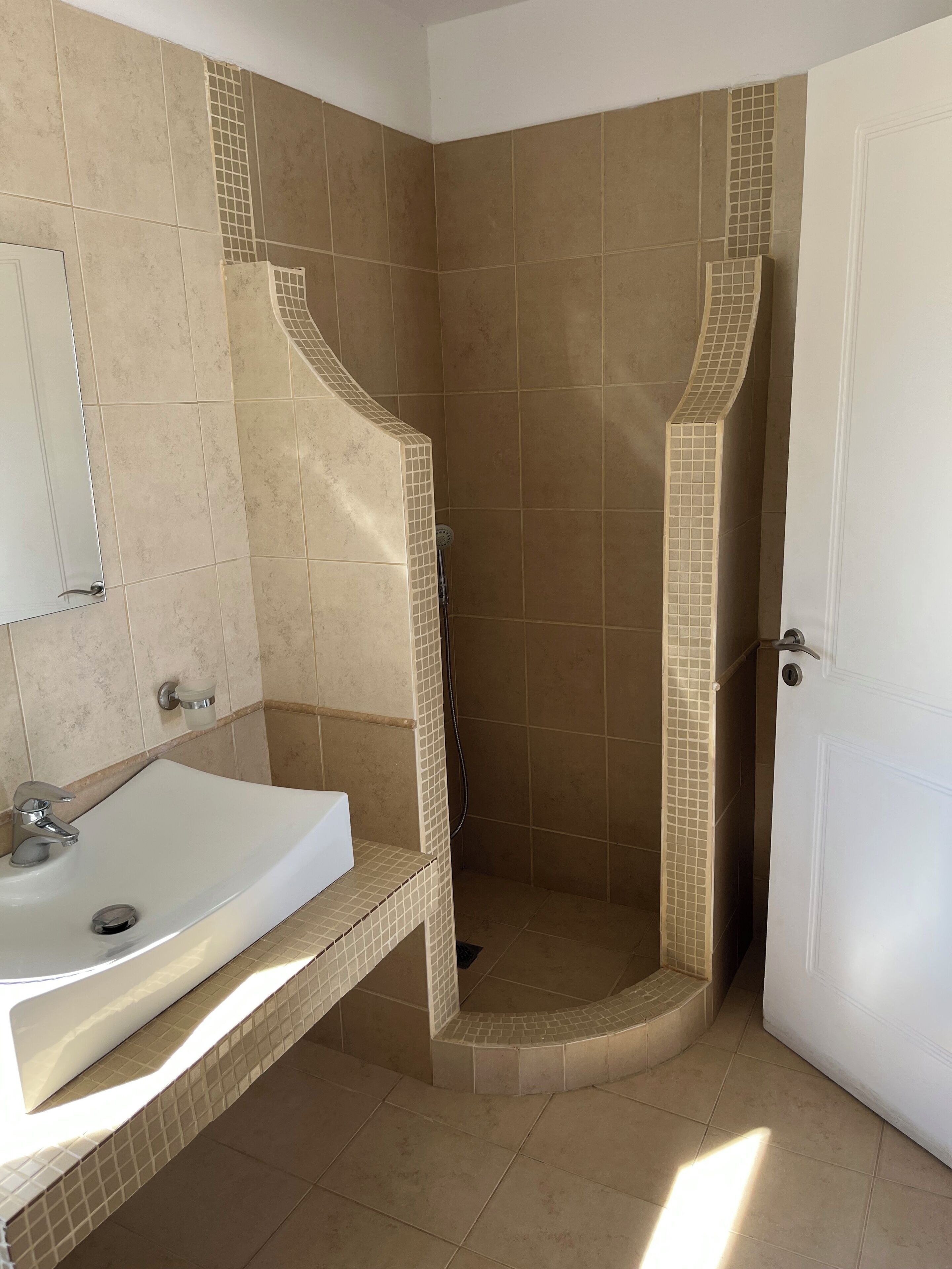 A bathroom with a white sink, a mirror, and a shower with beige tiles.