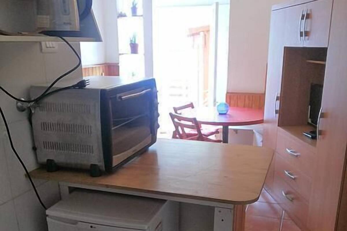A compact kitchen with a refrigerator, a microwave, and a small oven. There is a dining table with chairs, and a cabinet with drawers.
