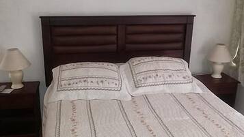 Une chambre à coucher avec une tête de lit en bois foncé, un couvre-lit blanc à motifs discrets, deux lampes de chevet et un luminaire suspendu.