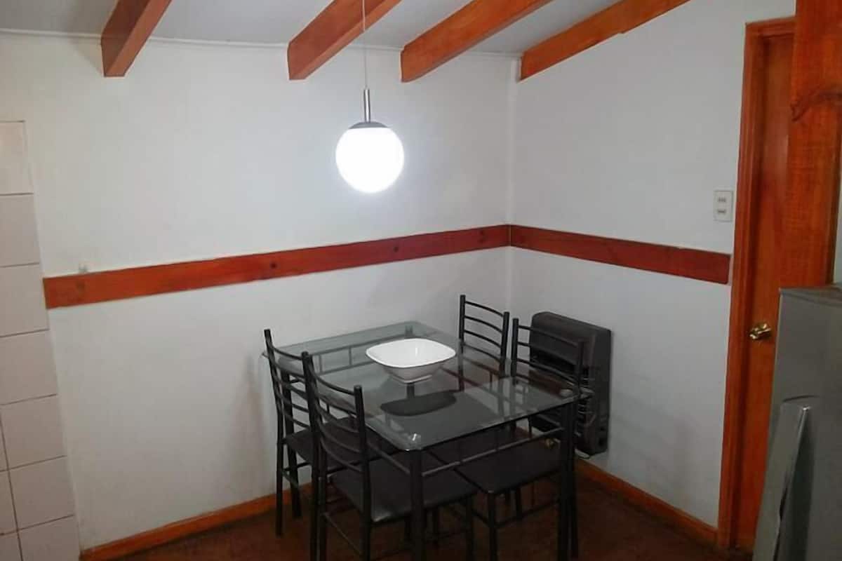 A small dining area with a glass table and black chairs, a single hanging light, and a wooden ceiling beam.
