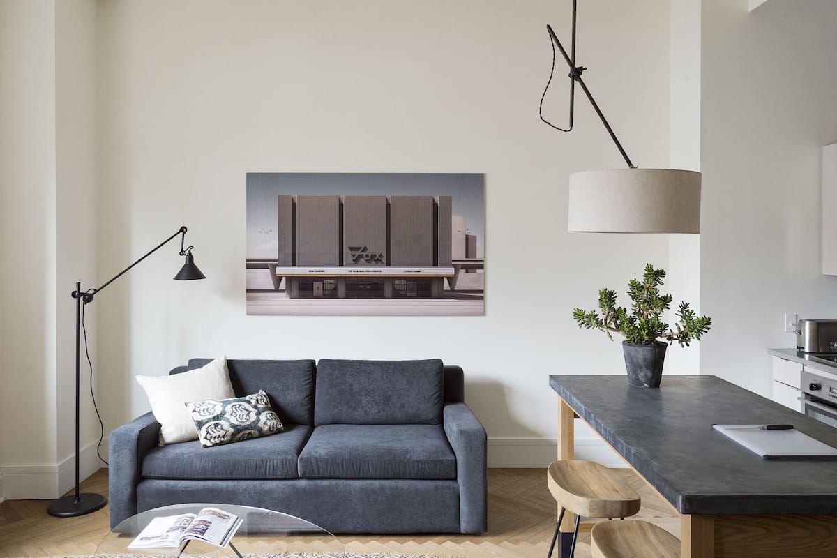 A modern living room with a grey sofa, a coffee table, a floor lamp, a dining table with chairs, a potted plant, and a framed picture on the wall.